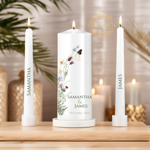 A beautifully designed wedding unity candle set, featuring a large central candle and two taper candles in white. The central candle is adorned with a delicate floral illustration with wildflowers and a butterfly, and includes an elegant serif font for personalization. The taper candles complement the design with matching typography. The set is displayed on a wooden surface with a warm, softly lit background featuring candles and stylish décor. The VG Designs IE logo is subtly placed in gold.